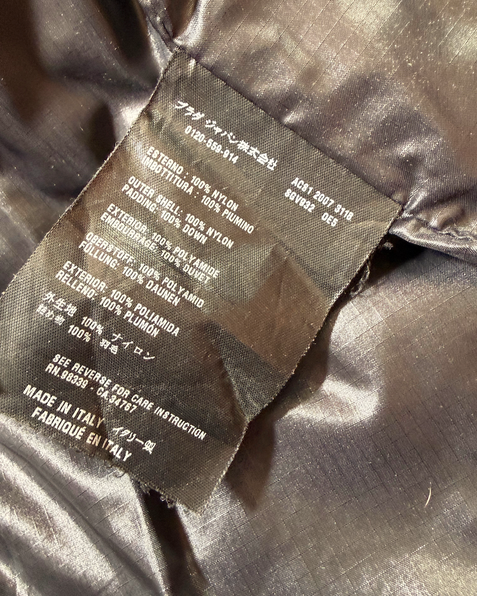 Close-up of a sewn-in product Prada label on shiny dark brown or bronze fabric, showing text including material composition and Made in Italy.