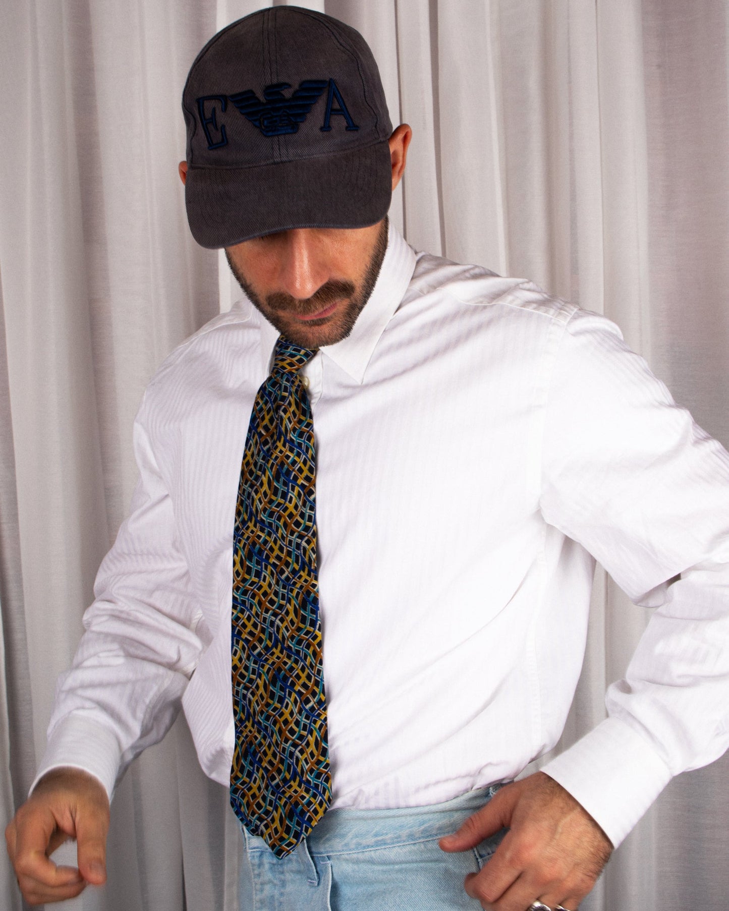 Man wearing a white shirt, patterned vintage missoni tie, and dark cap with logo against a plain background