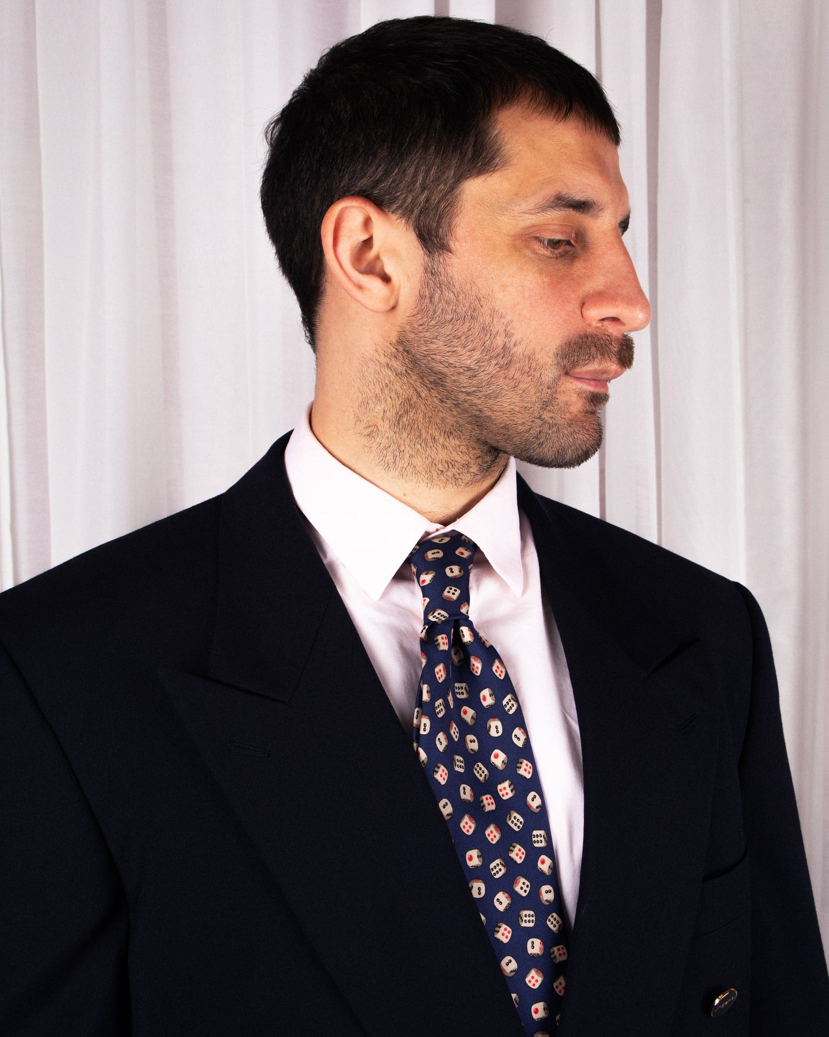Man wearing a dark suit and patterned gucci tie against a white curtain background