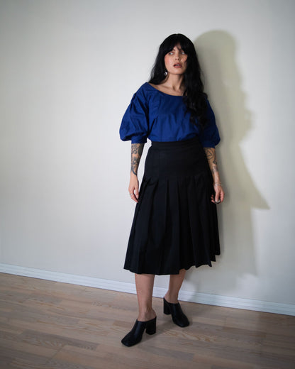 Woman wearing a blue top and black fendi skirt standing against a plain white wall.