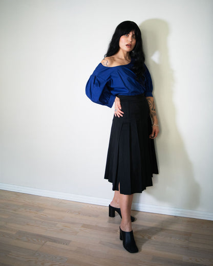 Woman wearing a blue top and black fendi skirt against a white wall.