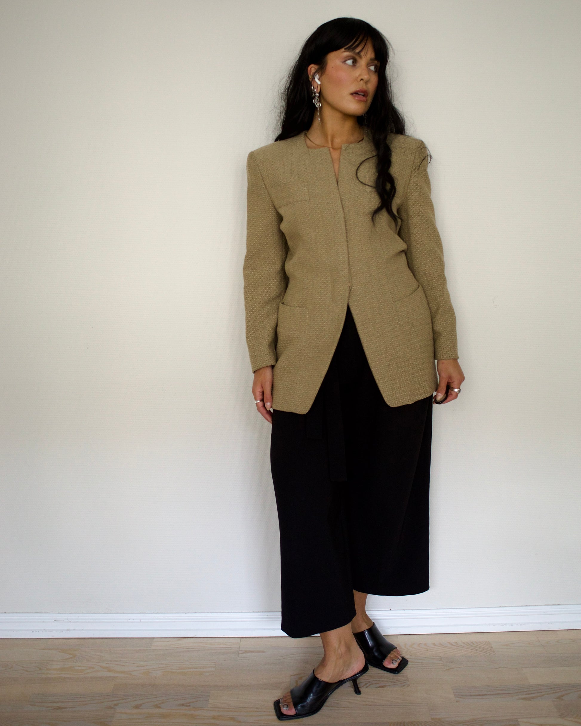 Woman wearing a beige blazer and black pants against a white wall.