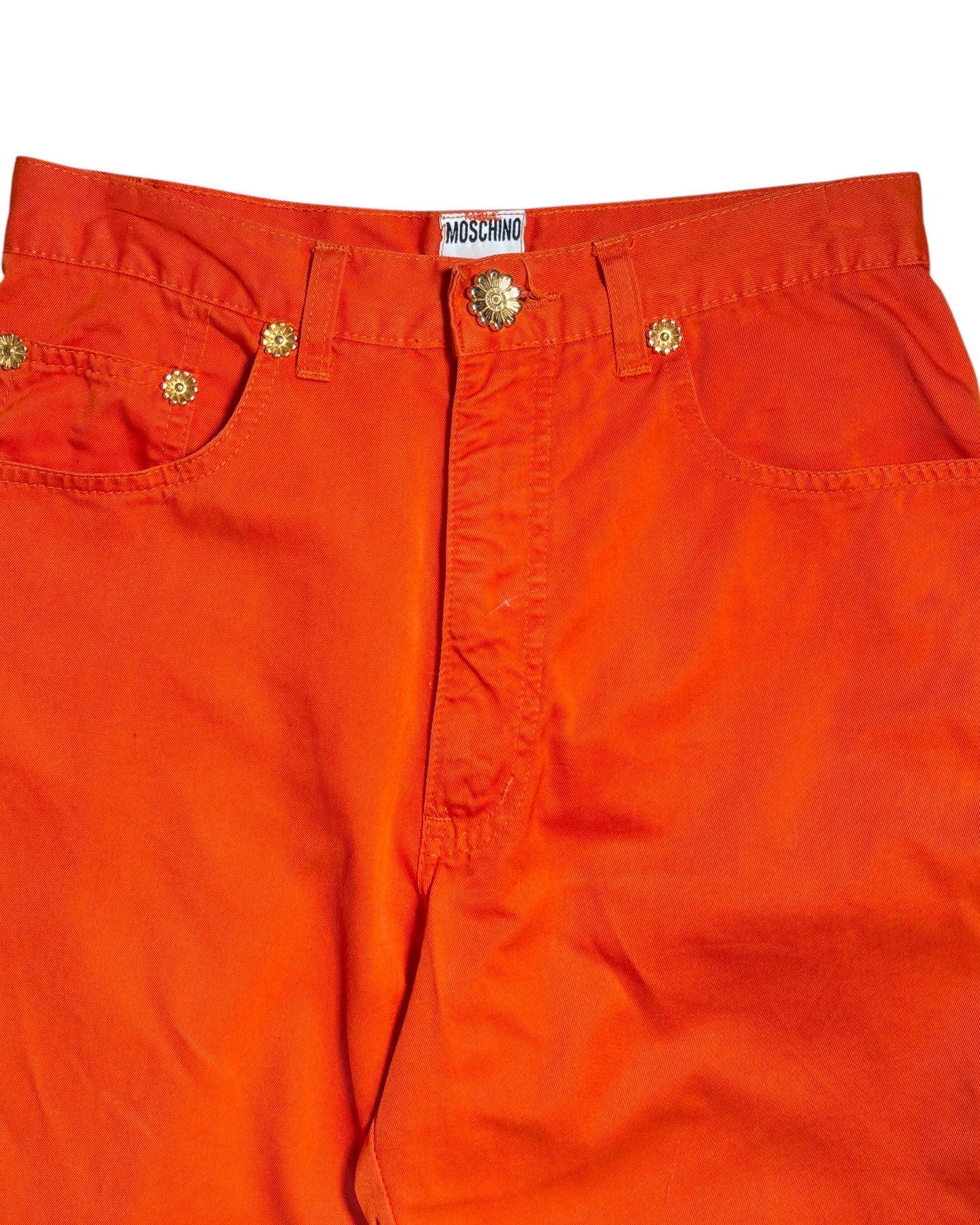 Bright orange denim pants featuring gold-tone metal rivets, a decorative button closure, and a visible Moschino brand label at the waistband.