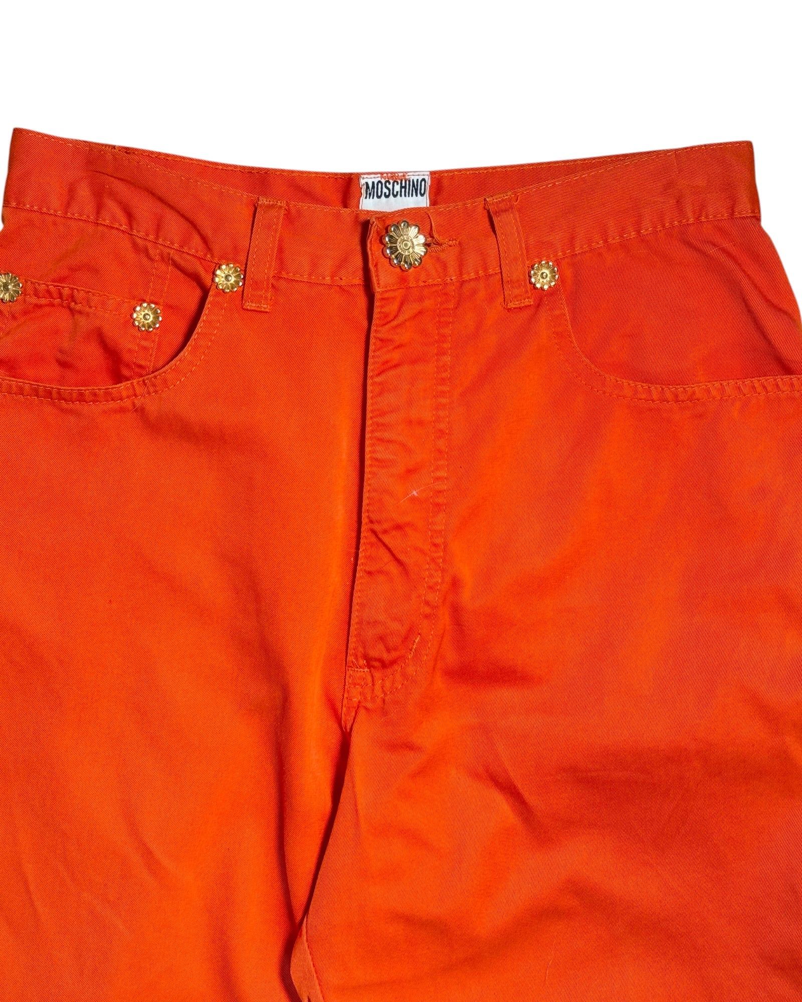 Bright orange denim pants featuring gold-tone metal rivets, a decorative button closure, and a visible Moschino brand label at the waistband.