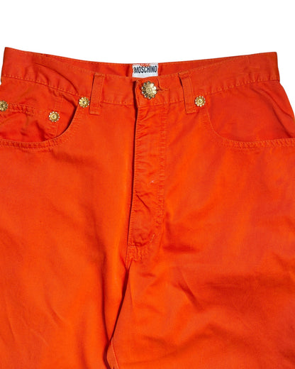 Bright orange denim pants featuring gold-tone metal rivets, a decorative button closure, and a visible Moschino brand label at the waistband.
