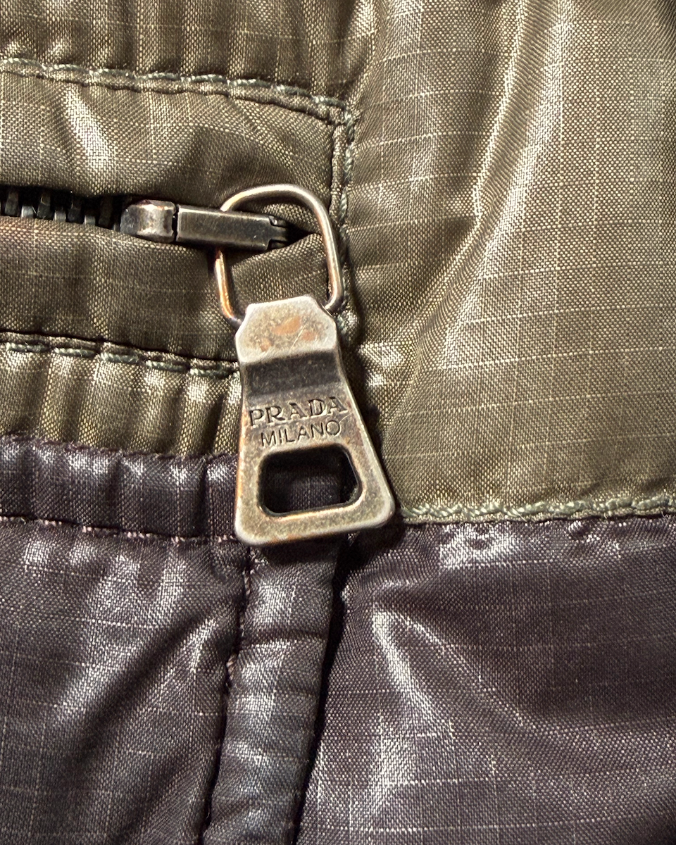 Close-up of a Prada zipper pull tab with 'PRADA MILANO' embossed on it, showing a textured olive green and dark brown nylon fabric.