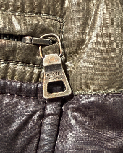 Close-up of a Prada zipper pull tab with 'PRADA MILANO' embossed on it, showing a textured olive green and dark brown nylon fabric.