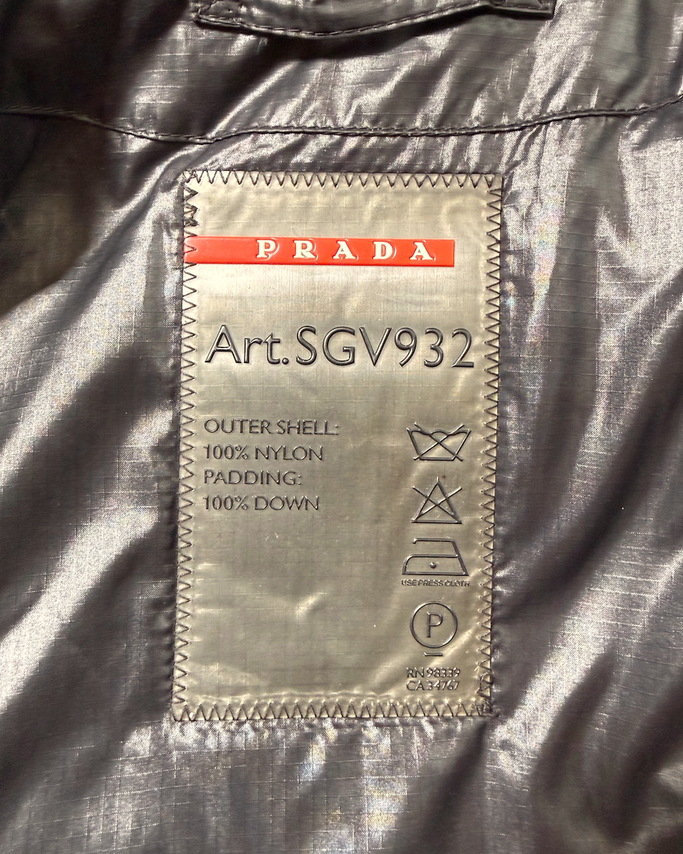 Close-up of a Prada garment interior label reading 'Art.SGV932' on a shiny silver-gray nylon fabric background.