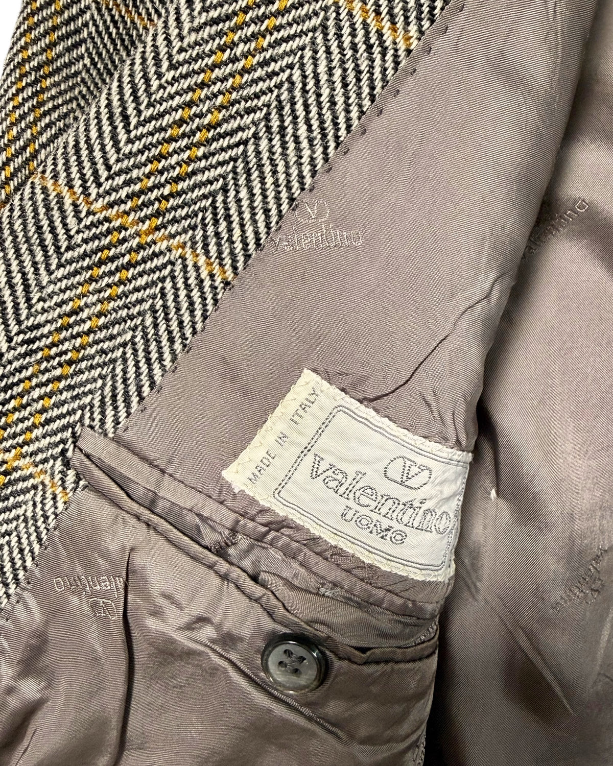 Close-up of a Valentino jacket showing the interior lining, a buttoned pocket, and a woven brand label. The lining is taupe-gray, and part of the outer fabric shows a black, white, and yellow herringbone pattern.