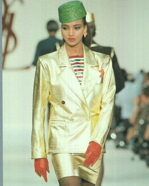 Model wearing a 1980s Yves Saint Laurent Gold Leather Jacket with a green hat and red gloves on a runway.